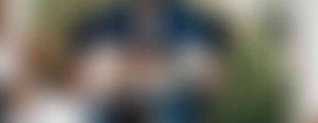 People kneel before a nativity scene in a church, surrounded by Christmas trees and lights. The mood is reverent and festive.