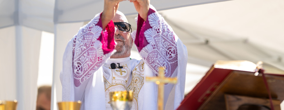 Priest in white and magenta vestments lifts a communion host at an outdoor Mass under a white canopy, with chalice and cross on altar.