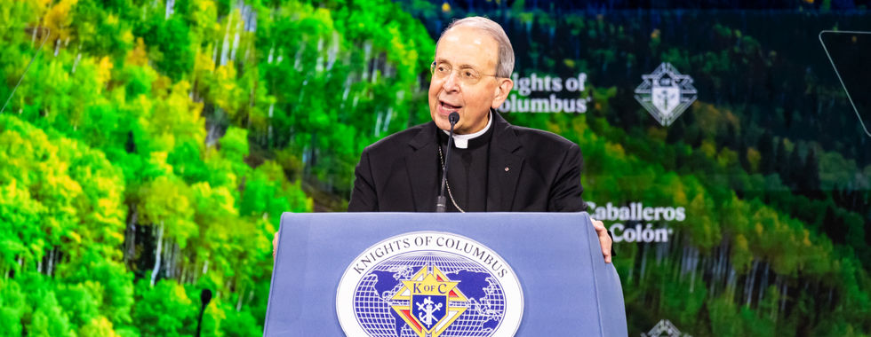 Catholic archbishop speaks at a Knights of Columbus Supreme Council podium before a green forest backdrop.