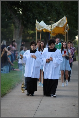 Two individuals in white robes participate in a procession, carrying incense and a canopy. Crowd along path; trees in background. Calm mood.