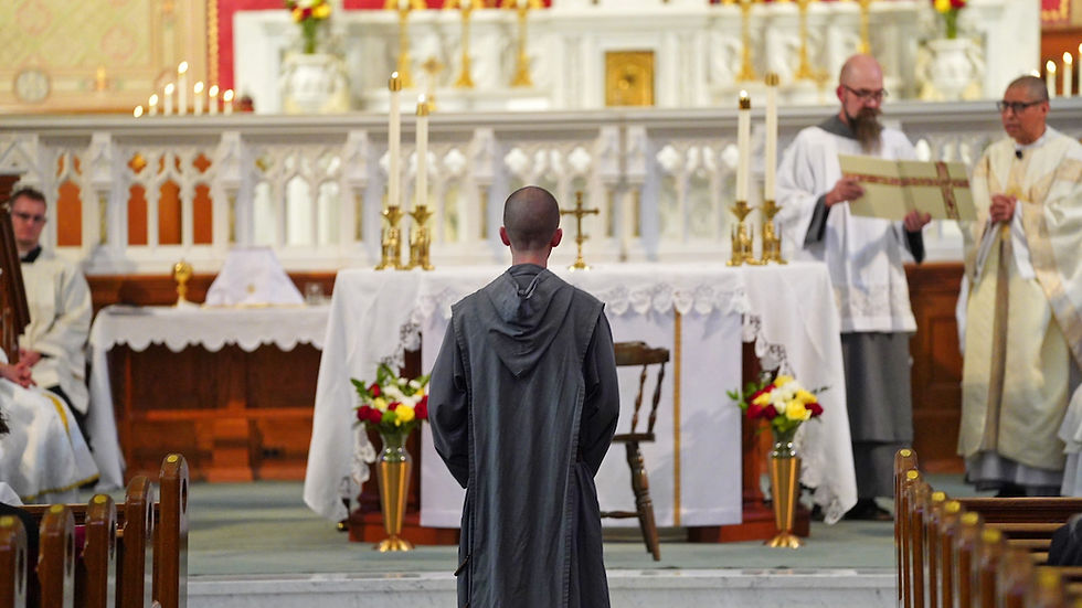 A monk in a gray robe stands before an altar in a church, with priests nearby carrying a scroll. The altar is adorned with candles and flowers.
