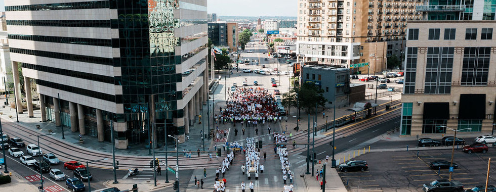Large march with a crowd filling a downtown street between tall buildings under a bright sky.