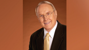 Smiling man in glasses wearing a dark suit and yellow tie against a warm brown background. Professional and welcoming mood.