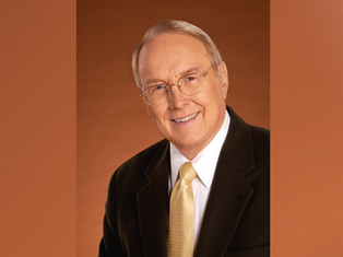 Smiling man in glasses wearing a dark suit and yellow tie against a warm brown background. Professional and welcoming mood.