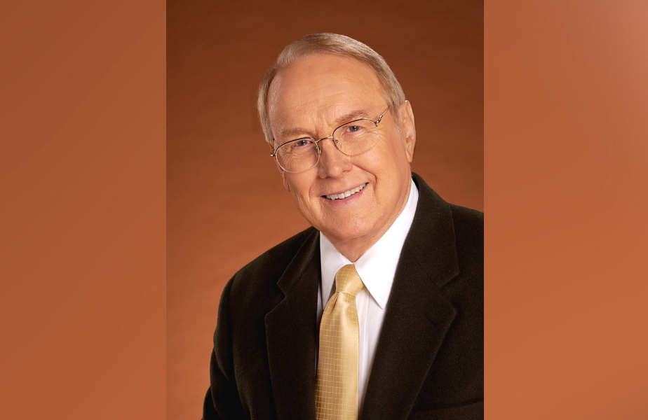 Smiling man in glasses wearing a dark suit and yellow tie against a warm brown background. Professional and welcoming mood.