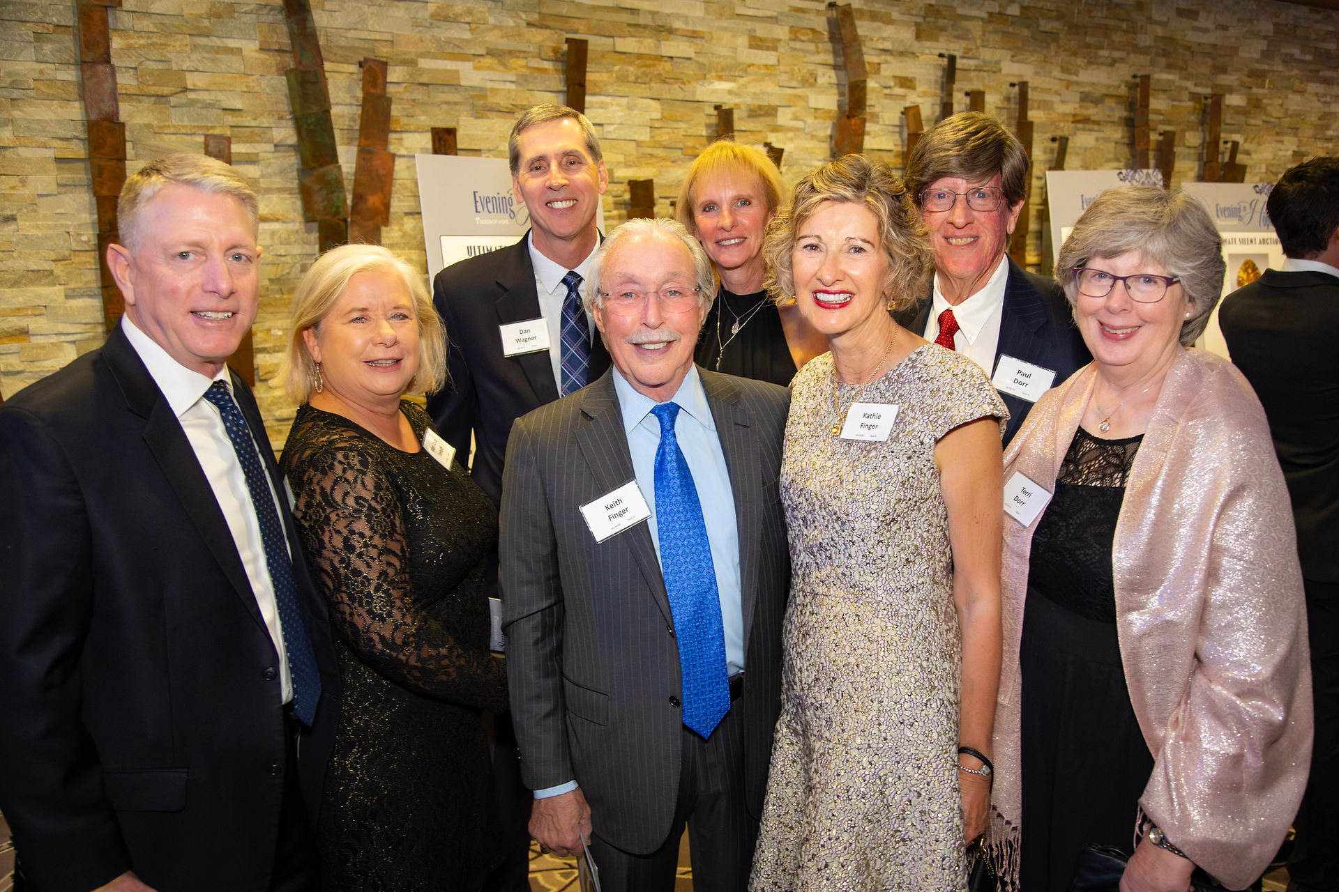 A group of elegantly dressed people smiling at an event. Stone wall background, name tags visible. Warm, celebratory atmosphere.