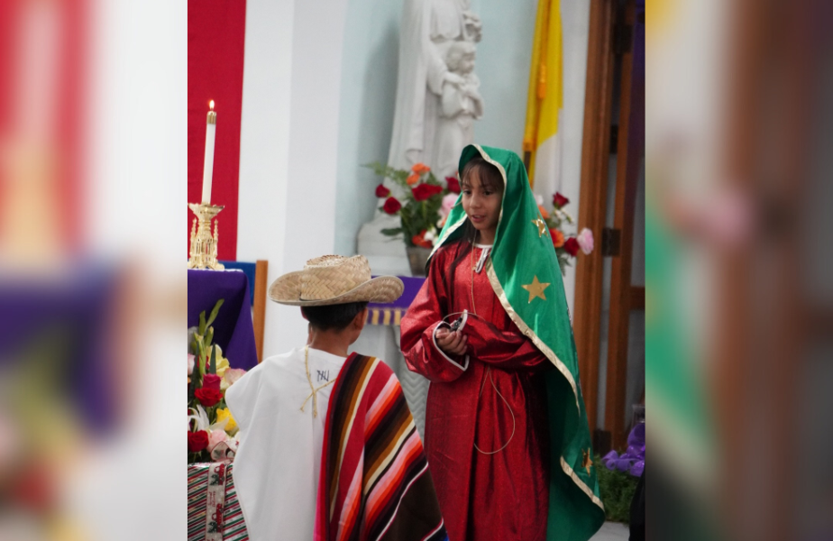 Children in traditional attire perform in a church. Girl in green and red, boy in sombrero. Candles, flowers, and statue in the background.