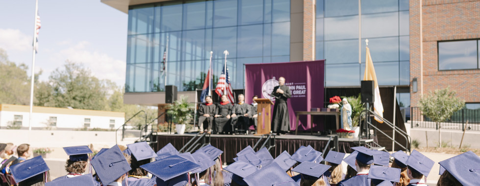 Graduates in blue caps watch an outdoor commencement stage with a speaker, flags, and a John Paul the Great banner by a modern building.
