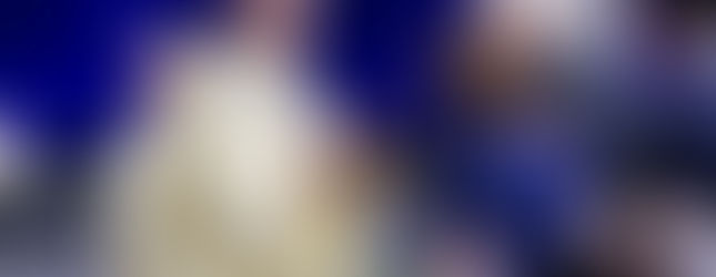 Bishop in gold vestments offers communion to bowed worshippers on a stage with blue curtains and a religious portrait