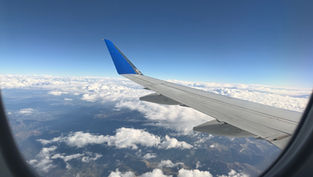 Airplane wing seen from a window above white clouds and mountains under a clear blue sky, calm and expansive view