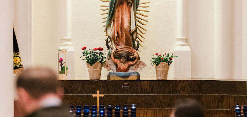 People praying in a church before a statue of Our Lady of Guadalupe. Lit candles and flowers adorn the altar, creating a serene atmosphere.