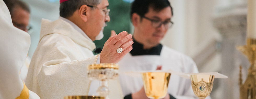 A bishop in a white robe and pink cap leads a religious ceremony with a priest, surrounded by golden chalices, in an ornate church setting.