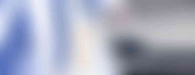 Clergy in white robes with sashes walk on a sunny pavement. One holds a rosary. Background is dark and shadowed. Peaceful mood.