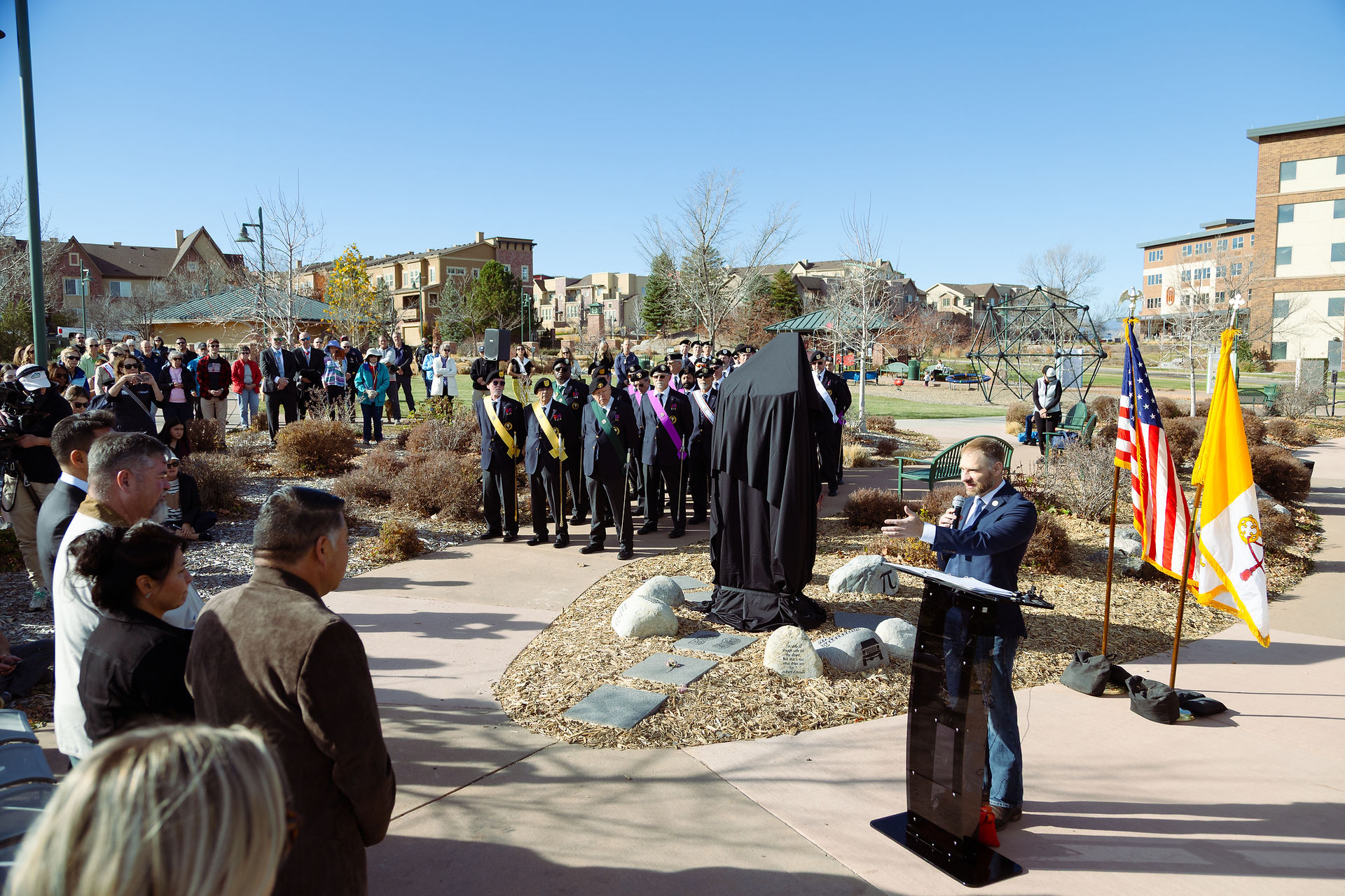 A speaker addresses a crowd at an outdoor ceremony beside flags and a draped statue. People watch attentively in a park setting under clear skies.