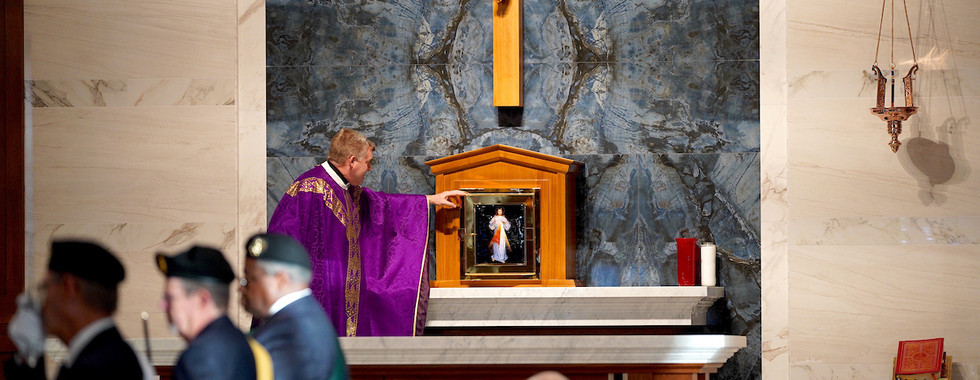 Priest in a purple robe points at an image on a church altar. Marble wall, crucifix above. People in foreground, solemn mood.