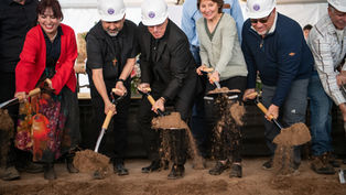 People in hard hats and colorful attire participate in a joyous groundbreaking ceremony, shoveling dirt indoors.