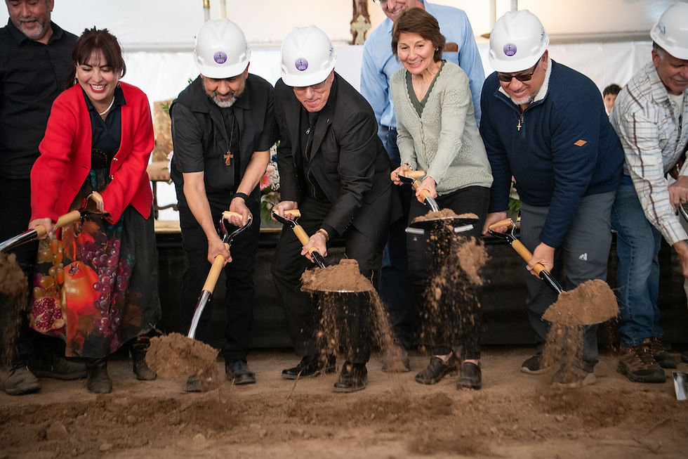 People in hard hats and colorful attire participate in a joyous groundbreaking ceremony, shoveling dirt indoors.