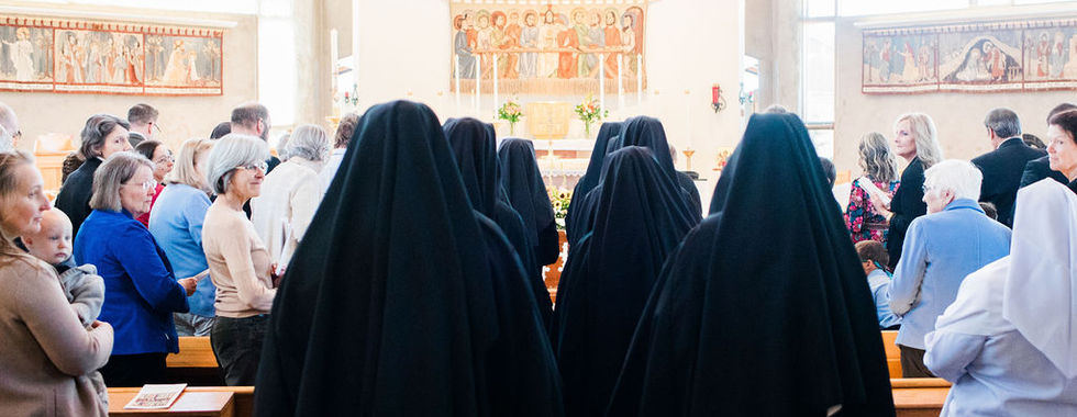 Religious sisters in black habits walk down the center aisle of a chapel as family, friends and fellow sisters gather for a profession of vows ceremony.