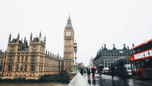 View of Big Ben and the Houses of Parliament in London. Red double-decker buses and people with umbrellas on wet street. Overcast sky.