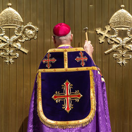 A person in religious attire, holding a hammer, faces a gold door with ornate emblems, wearing a purple and red embroidered robe.