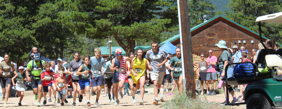 Group of runners, including children and adults with bibs, races on a dusty forest road beside a cabin under pine trees and mountains