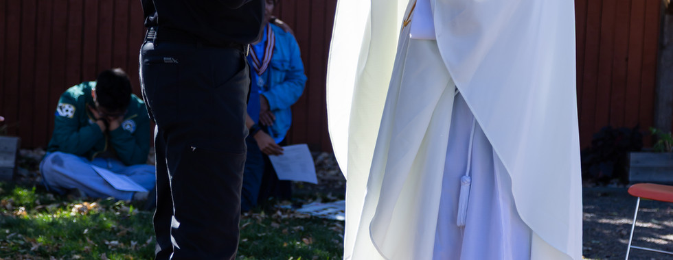Priest in white vestment holds up Eucharist outdoors, facing man in black. Green grass, trees, and a wooden fence in the background.