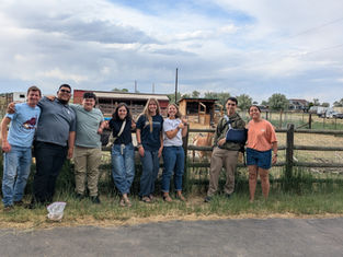 Smiling group of eight stands by a wooden fence at a farm, with barns, pens, and a cloudy sky behind them.