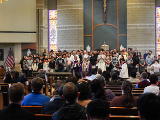 Church ceremony with clergy in purple robes and diverse group of people standing, stained glass windows behind, congregation seated.