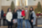Family of six poses inside a decorated church with Christmas trees and wreaths. A child in red adds a festive touch. Smiling faces.