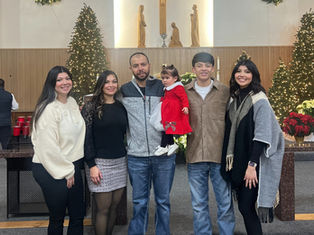 Family of six poses inside a decorated church with Christmas trees and wreaths. A child in red adds a festive touch. Smiling faces.
