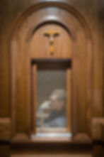 Man in confessional, viewed through lattice, in wooden booth adorned with crucifix. Dim lighting, solemn atmosphere.