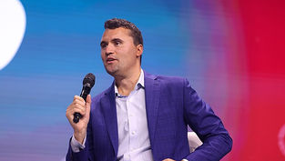 Man in a blue suit holding a microphone on stage, speaking. Background is a blurred gradient of blue and red, conveying a focused mood.
