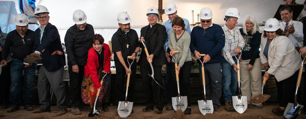 Clergy and community members wearing hard hats participate in a ceremonial groundbreaking with shovels.