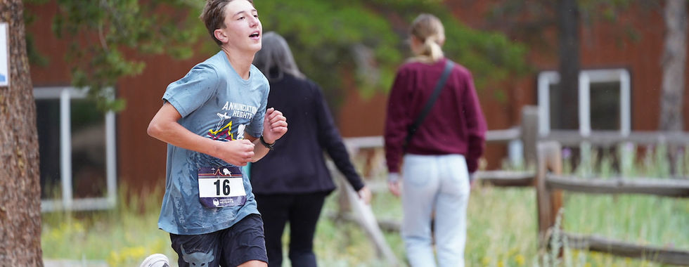 Teen boy running in a race, wearing number 16 and a blue shirt. Background: two people walking, wooden fence, green foliage. Energetic mood.