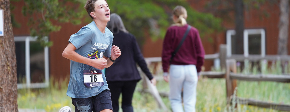 Teen boy running in a race, wearing number 16 and a blue shirt. Background: two people walking, wooden fence, green foliage. Energetic mood.