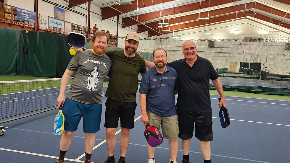 Four men smiling on an indoor tennis court, each holding a pickleball paddle. Background shows nets and banners. Casual mood.