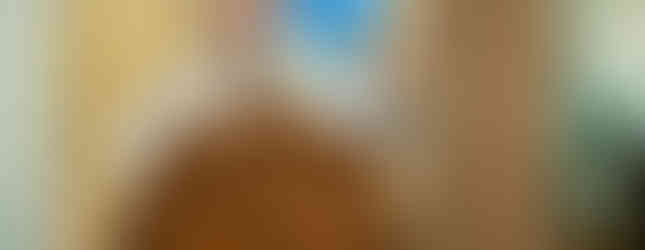 A priest in ornate robes speaks at a wooden pulpit with a cross, gesturing with one hand. Blurred religious painting in the background.