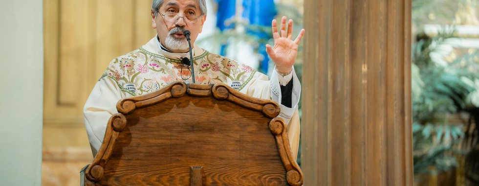 A priest in ornate robes speaks at a wooden pulpit with a cross, gesturing with one hand. Blurred religious painting in the background.