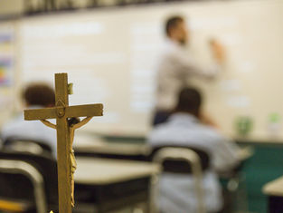 Crucifix in focus in a classroom; blurred teacher writes on whiteboard. Students seated, vibrant wall colors in the background.
