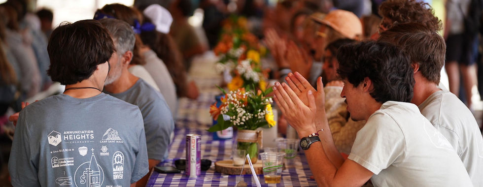 People seated at a long table with a blue checkered tablecloth, clapping. Visible flowers and text on shirts. Lively, communal atmosphere.