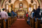 People standing in church pews during a service, facing an ornate altar with statues and floral decorations. The mood is solemn.
