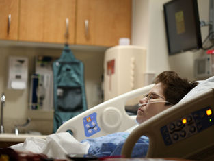 Person in a hospital bed with oxygen tube, eyes closed. Medical equipment and cabinets in the background. Calm, clinical setting.