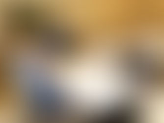 Five elderly men smiling around a dining table with plates of food and drinks. One stands, while others sit. A framed painting decorates the wall.