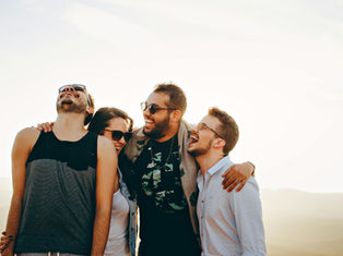 Four friends laugh together outdoors, wearing casual clothes and sunglasses, against a bright sky. Warm, joyful mood.