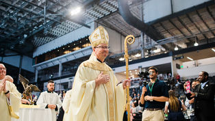 A bishop in ornate robes and hat holds a staff in a large indoor venue, surrounded by clergymen, photographers, and attendees.