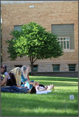 People sit on grass in a park setting, near a tree and brick building, enjoying a sunny day. They appear relaxed and engaged in conversation.