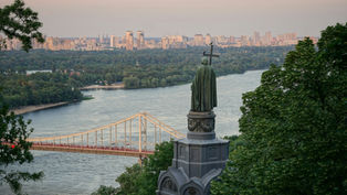 Statue on a hill overlooks a river, bridge, and distant city skyline framed by trees at sunset.