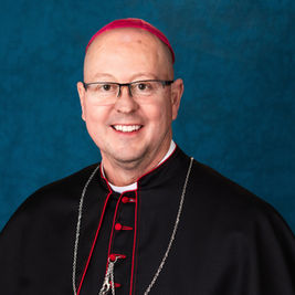Hombre sonriente con sotana negra, solideo fucsia y collar, posando frente a fondo azul. Ambiente formal y sereno.