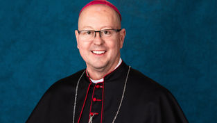 Hombre sonriente con sotana negra, solideo fucsia y collar, posando frente a fondo azul. Ambiente formal y sereno.