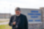 Bishop in black holds a rosary outside Hudson Correctional Facility sign in Hudson, Colorado, looking somber.